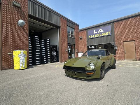 View of the garage's parking lot, with the open bay, stacked tires, and a green sports car parked out front.