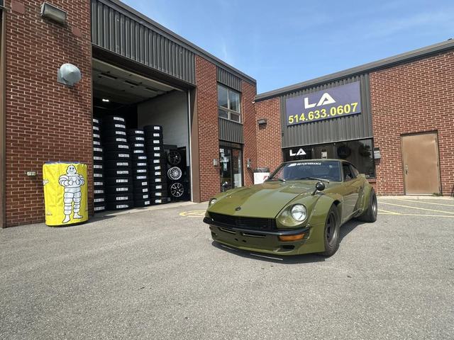 View of the garage's parking lot, with the open bay, stacked tires, and a green sports car parked out front.